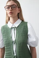 Chaleco tejido de punto color beige con trama texturada y diseño liviano. Presenta cuello a la base, silueta recta y cierre frontal mediante tres lazos delicados.