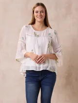 Blusa blanca de gasa con bordados florales, corte holgado, mangas 3/4 acampanadas con detalle de puntilla y forro interno.