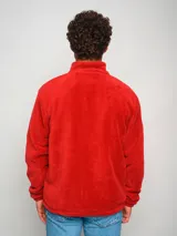 Campera polar roja con cierre frontal, cuello alto y bolsillos laterales. Presenta un panel liso en la parte superior del pecho y un bolsillo con cierre en el lado izquierdo. Incluye el logo de Umbro bordado en el pecho izquierdo y el escudo del Club Nacional de Football bordado en el pecho derecho.