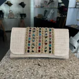 Clutch tipo sobre de fiesta, confeccionado en tejido color natural con bordado artesanal de piedras de vidrio multicolores. Incluye asa de cadena dorada para colgar al hombro.
