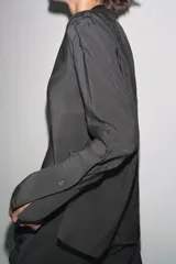 Camisa negra de lyocell con cuello redondo, manga larga con puños y bajo asimétrico con aberturas laterales. Cierre frontal con botones.