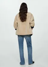 Chaqueta beige de poliéster con textura de borreguito, cuello redondo, cierre frontal con botones tipo trenca, ribetes en contraste de color negro, dos bolsillos delanteros y forro interior.
