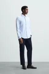 Camisa celeste a rayas verticales blancas, de corte recto, con cuello italiano abierto, mangas largas con puños abotonados y cierre frontal con botones.