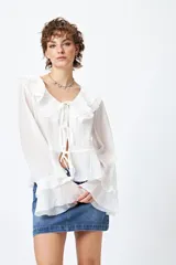 Blusa blanca de gasa con escote en V y volados en el cuello y las mangas largas. Se ata con lazos finos al frente.
