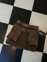 Falda corta de cuero gamuza color marrón oscuro, con diseño utilitario que incluye grandes bolsillos frontales con solapa y un anillo metálico en la cintura. Incluye un cinturón removible a juego.