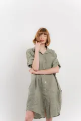 Vestido camisero corto color verde oliva, confeccionado en lino 100%. Tiene botones en la parte delantera, cuello camisero, mangas cortas con botón para arremangar y bolsillos laterales.
