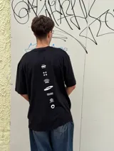 Remera negra de corte oversize con cuello redondo y manga corta. Presenta tres estampados blancos en el frente: un símbolo curvo a la izquierda, la palabra "OSTEN" en el centro y un símbolo abstracto a la derecha.