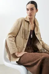 Chaqueta color beige de simil gamuza con flecos en el frente y en la espalda. Tiene cuello clásico, cierre con botones y bolsillos delanteros.