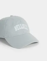 Gorra deportiva color crudo, de tela canvas con bordado frontal de la palabra "Wellness" en letras negras estilo universitario. Tiene visera curva y tira ajustable trasera con hebilla metálica.