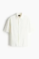 Camisa de manga corta color crema, de corte holgado, con cuello inglés, tapeta lisa, canesú en la espalda y bajo recto. Confeccionada en mezcla liviana de lino y viscosa.