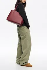 Bolso shopper de piel color borgoña, con textura granulada, asas de hombro y cierre de cordón. Presenta el logo metálico de Bimba y Lola en el frente.