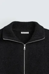 Cárdigan negro de punto con cuello alto, cierre frontal con cremallera, bolsillos laterales y mangas largas.