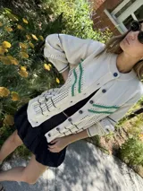 Saco de punto calado color beige claro, con cuello redondo y cierre frontal de botones azules. Presenta detalles decorativos en crochet verde y negro en el frente y puños.