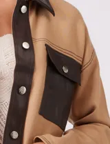 Chaqueta de corte boxy confeccionada en tejido de punto color crema, con detalles en cuero sintético negro en cuello, solapas, cartera central y bolsillos frontales. Presenta cierre frontal con botones metálicos plateados y costuras decorativas visibles.