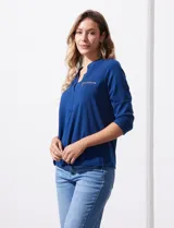 Blusa blanca de poliéster y elastano, con cuello a la base, escote en V y mangas tres cuartos. Presenta dos bolsillos frontales con cierre metálico dorado.