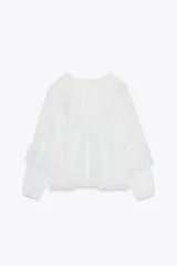 Blusa blanca semitransparente de escote pico y manga larga con puño elástico. Detalle de volantes y bordados a tono. Cierre frontal con botones.
