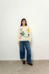 Sweater de lana color crudo con cuello redondo y mangas largas. Presenta apliques de tela cosidos con forma de flores y figuras geométricas en tonos gris, azul, verde, amarillo y rosa.