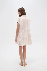 Vestido corto con cuello camisero, de corte holgado y mangas cortas. Presenta un diseño a rayas verticales en tonos beige y blanco, con volados en la falda y cierre frontal con botones.