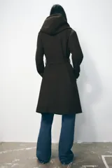 Abrigo largo color beige con capucha, cuello con solapa, manga larga, bolsillos laterales y cinturón lazada en el mismo tejido. Cierre frontal cruzado con botones.