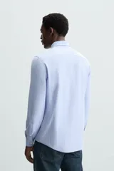 Camisa celeste a rayas verticales blancas, de corte recto, con cuello italiano abierto, mangas largas con puños abotonados y cierre frontal con botones.