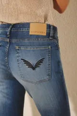 Jean de mujer color azul oscuro, tiro alto, corte oxford (o campana moderada) con desgaste localizado y bordado de alas negras en los bolsillos traseros.