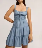 Vestido corto color beige, con escote recto, breteles medianos y lazo decorativo en el centro del busto. Presenta costura fruncida en la cintura y falda con babados en capas.