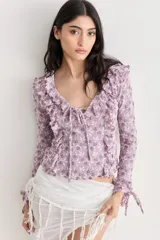 Top de encaje con estampado floral en tonos lila y blanco, escote en V con volados y lazos ajustables, mangas largas con puños elásticos y volados.