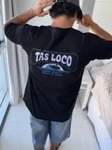Remera negra de corte holgado con estampado gráfico en la espalda que dice "TAS LOCO" en letras rosas y celestes, con una ilustración de una ola y el texto "SUNNY WITH A CHANCE OF STOKE".