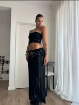Conjunto de dos piezas negro compuesto por un top tipo bandeau y una falda larga de tejido de punto calado. El conjunto se complementa con un cinturón ancho de cuero negro con tachas metálicas y hebilla cuadrada.