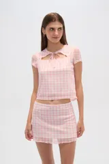 Conjunto de top y falda a cuadros vichy en tonos rosa y blanco. El top es de manga corta, corte crop y escote cuadrado con abertura y lazo. La falda es corta, ajustada y con volantes en el ruedo.