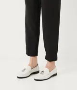 Zapato mocasín de color blanco con textura de piel de cocodrilo y detalle de borla frontal. Posee punta redonda, tacón plano y suela en contraste de color negro.