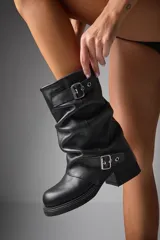 Bota negra de caña media, estilo motociclista, con efecto arrugado (slouchy) y hebillas decorativas en el empeine y en la caña. Posee suela de plataforma gruesa y taco bajo.