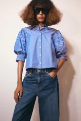 Camisa de cuello redondo con estampado de rayas verticales azules y blancas, manga larga abullonada y cierre frontal con botones.