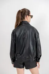Chaqueta bomber de cuero ovino color marrón oscuro, con cuello clásico, hombros caídos y ajuste elástico en la cintura. Presenta bolsillos con vivos y broches ocultos, y ajustes metálicos en puños y espalda.