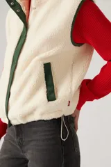 Chaleco color crema de sherpa con cuello alto, cierre frontal con cremallera y ribete de contraste en verde.