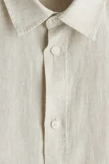 Camisa de lino color crema, de corte recto, con cuello inglés, tapeta clásica, mangas largas con puños abotonados y bajo redondeado.