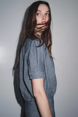 Camisa de denim azul con cuello redondo, manga corta abullonada con pliegues y bajo abullonado con pliegues. Cierre frontal con botones ocultos por solapa.