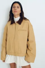 Campera color beige de algodón Oxford con cuello de pana marrón en contraste. Modelo de corte holgado con cierre frontal, hombros caídos, bolsillos laterales ribeteados y bolsillo superior con cierre. Ajuste con botones a presión en los puños y en la parte posterior del bajo.