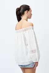 Blusa blanca de algodón con escote off-shoulder, mangas largas con puños elásticos y detalles de bordado y puntillas en escote y mangas.