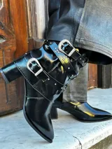 Botas de cuero sintético negro con punta fina, caña media con correas y hebillas plateadas, y taco cuadrado.