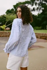 Blusa celeste con rayas verticales blancas, cuello redondo con escote en pico y mangas largas abullonadas terminadas en puño elástico. Presenta detalle de tejido en relieve con forma de flor.