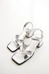 Sandalias plateadas de cuero, con tiras cruzadas sobre el empeine, pulsera al tobillo con hebilla y taco bajo cuadrado.