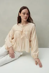 Blusa color beige de caída fluida con cuello redondo y lazos frontales. Diseño con cortes horizontales y frunces, mangas largas con puños elastizados.