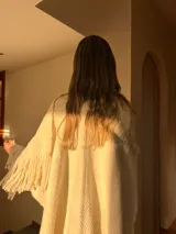 Ruana color crema tejida en punto grueso con cuello alto y flecos en el borde inferior.