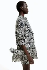 Vestido corto de algodón con estampado abstracto en blanco y negro. Tiene cuello con volados y lazo con borlas, mangas largas abullonadas con puños elásticos y falda escalonada con volados.