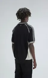 Chomba tejida de hilo color negro con punto cerrado, cuello polo con escote en V y ribetes blancos en cuello y mangas. Presenta bordados en blanco: un número "90" y la palabra "Ravers" en cursiva.