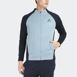 Campera deportiva con cierre frontal, cuello alto y mangas raglán. El cuerpo es de color celeste claro con textura acanalada, y las mangas y paneles laterales son de color azul marino oscuro. Presenta el logo de Le Coq Sportif bordado en el pecho.