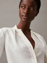 Blusa blanca de poliéster reciclado con escote en V, manga larga y detalle de pliegues en los hombros.