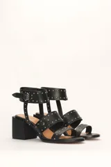 Sandalia de cuero vacuno color negro con tachas plateadas, tira ancha sobre los dedos, pulsera al tobillo con hebilla y taco cuadrado.