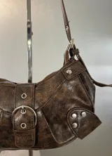 Cartera marrón con textura de cuero, forma de media luna y correa de hombro ajustable. Tiene cierre superior con cremallera y un bolsillo frontal con hebilla decorativa.
